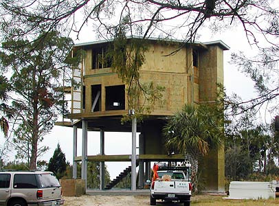 Florida Code-Compliant Elevated Home on Concrete Pilings: Hurricane-Resistant Design with Panoramic Views and Storm Surge Protection Elevated hurricane-resistant coastal home on concrete pilings under construction in Florida, compliant with strict building codes for storm surge and flood protection, offering panoramic views from all floors with open lower level, stairs, and surrounding pine trees.