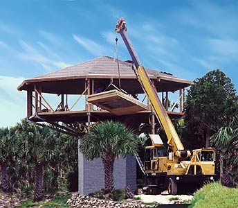 Crane lifting roof onto elevated 20-ft high oceanfront pedestal beach house during construction, hurricane-resistant design with palm trees and sandy site for storm surge and flood protection.