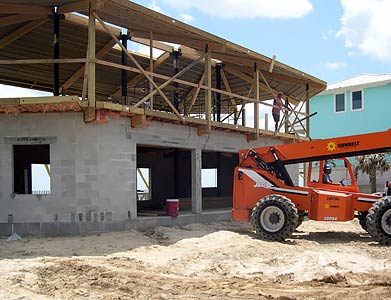 Elevated hurricane-proof beach house construction with reinforced concrete block foundation and flood vents for floodwater flow, featuring telescopic handler and open garage level on sandy coastal site.